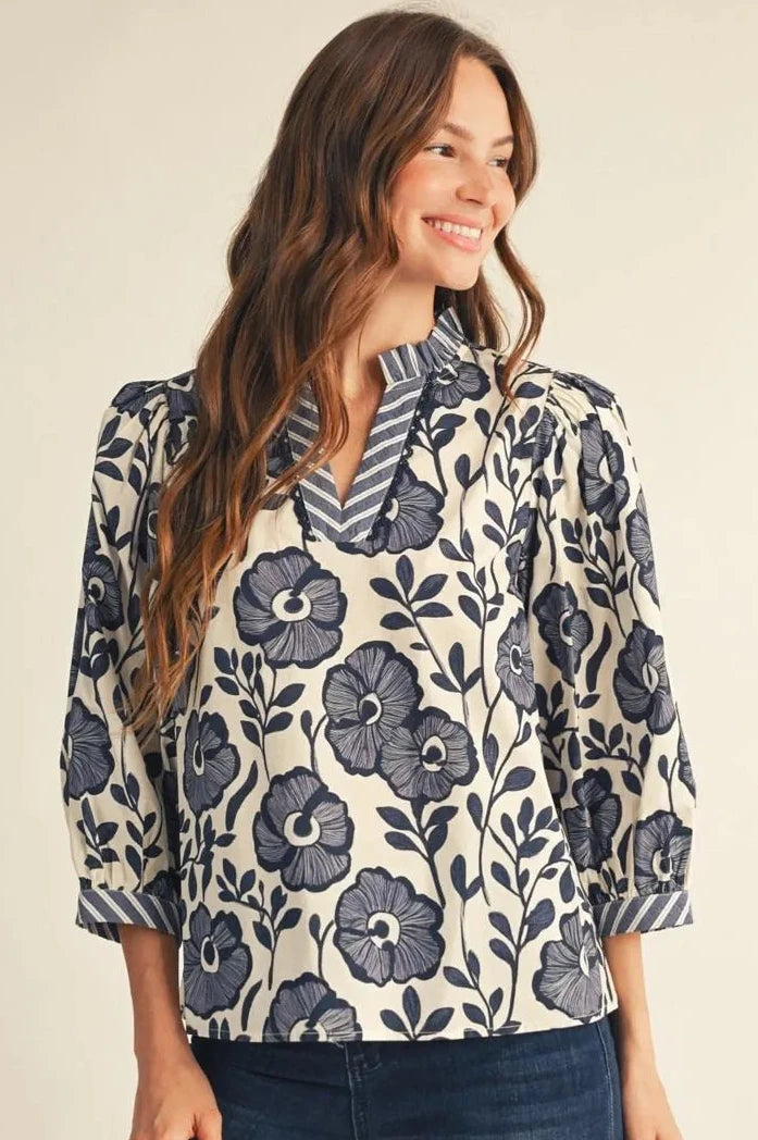 A woman wearing a cream-colored blouse with a navy blue floral and leaf pattern, paired with dark blue jeans, photographed against a solid light beige background.