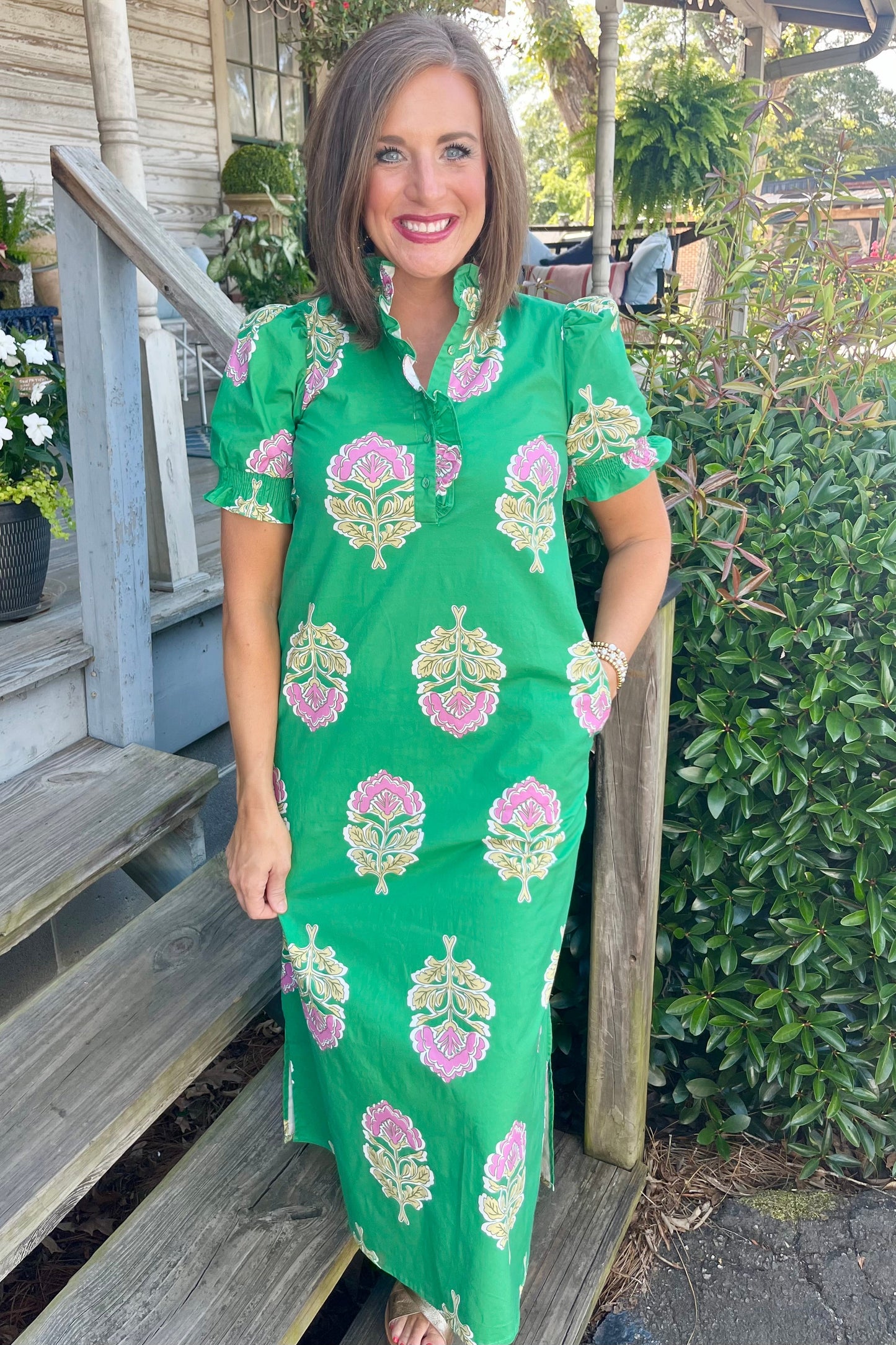 A woman wearing a bright green short-sleeve dress with pink and yellow floral block prints, standing on wooden porch steps outdoors.