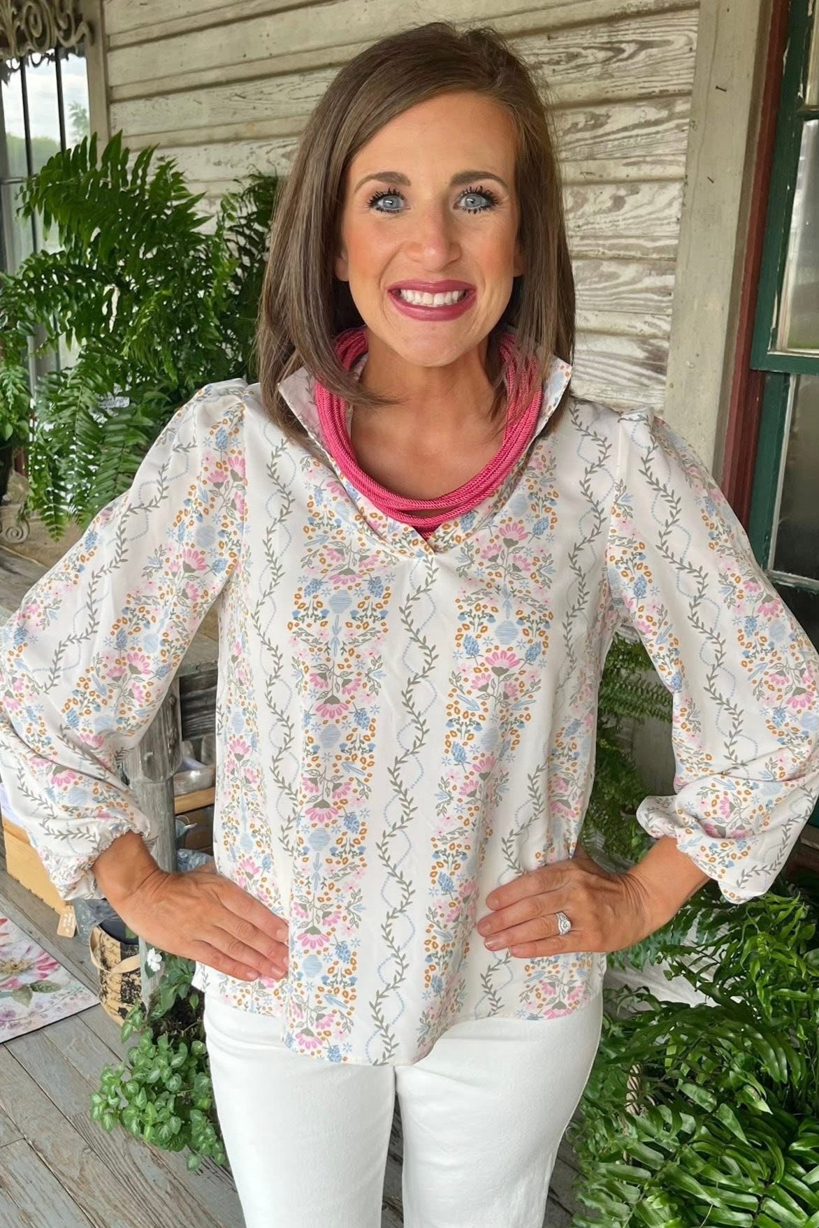 Woman wearing a floral blouse and white pants on a porch with plants and furniture.