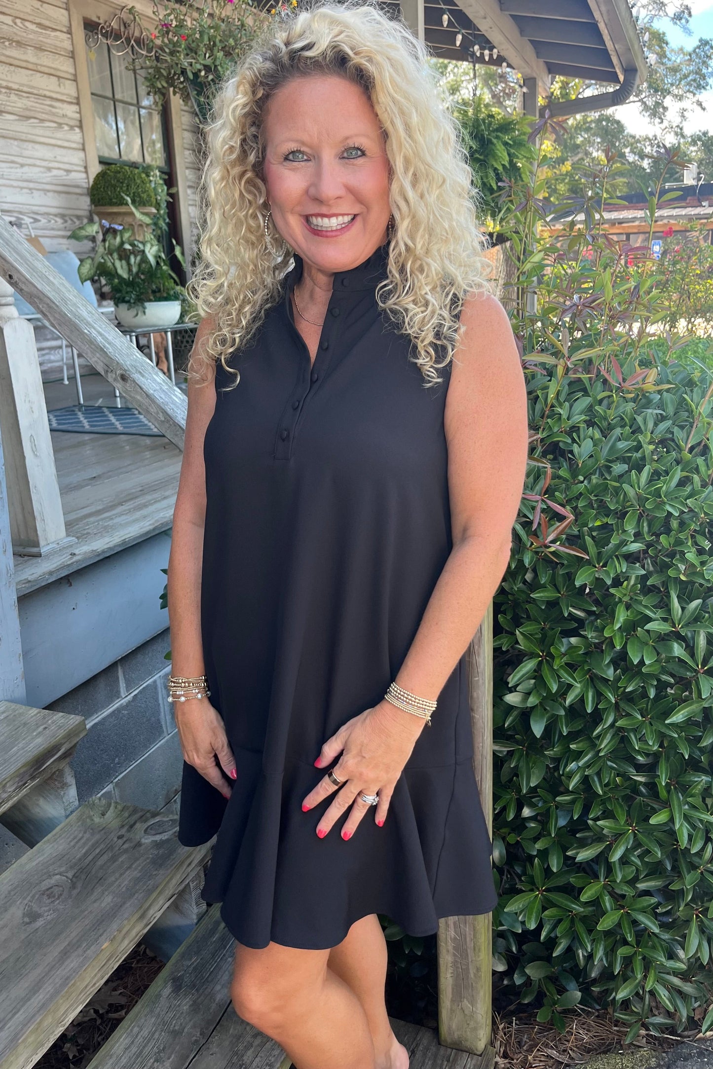 A smiling blonde woman wearing a sleeveless black dress with a high mock neck and flared skirt, standing on outdoor wooden steps next to a house and green bushes.