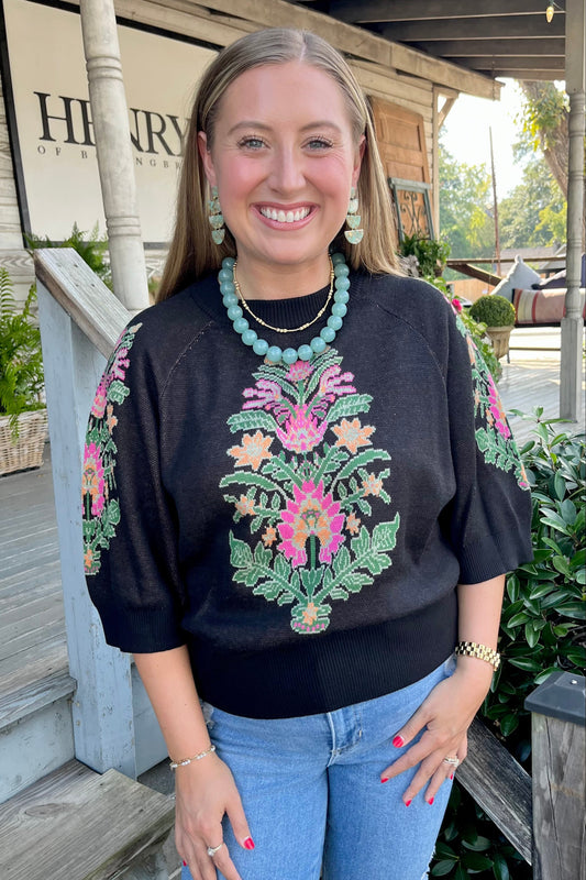 A smiling woman stands on a wooden porch wearing a black crewneck sweater with colorful floral embroidery on the front and sleeves, paired with light blue jeans. She is accessorized with a turquoise beaded necklace and matching earrings.