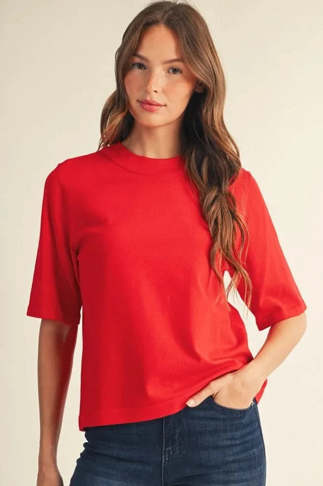 A woman wearing a bright red short-sleeve crewneck top and dark blue jeans, standing against a plain light beige studio background.