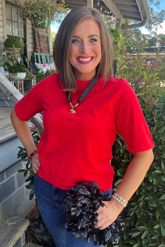 A woman smiling on a rustic outdoor porch wearing a bright red short-sleeve crewneck top, blue jeans, and holding a dark ruffled clutch.