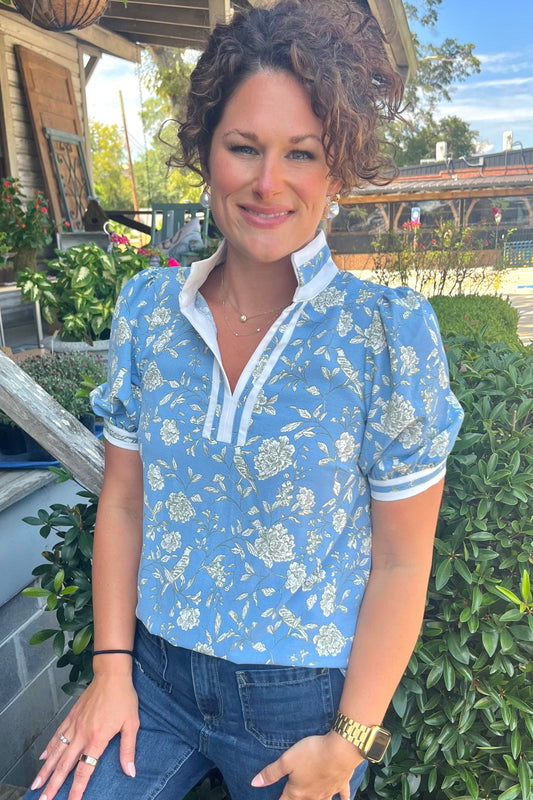 A woman with curly brown hair wearing a light blue and white floral puff-sleeve blouse and blue jeans, posing outdoors on a wooden porch with green bushes in the background.