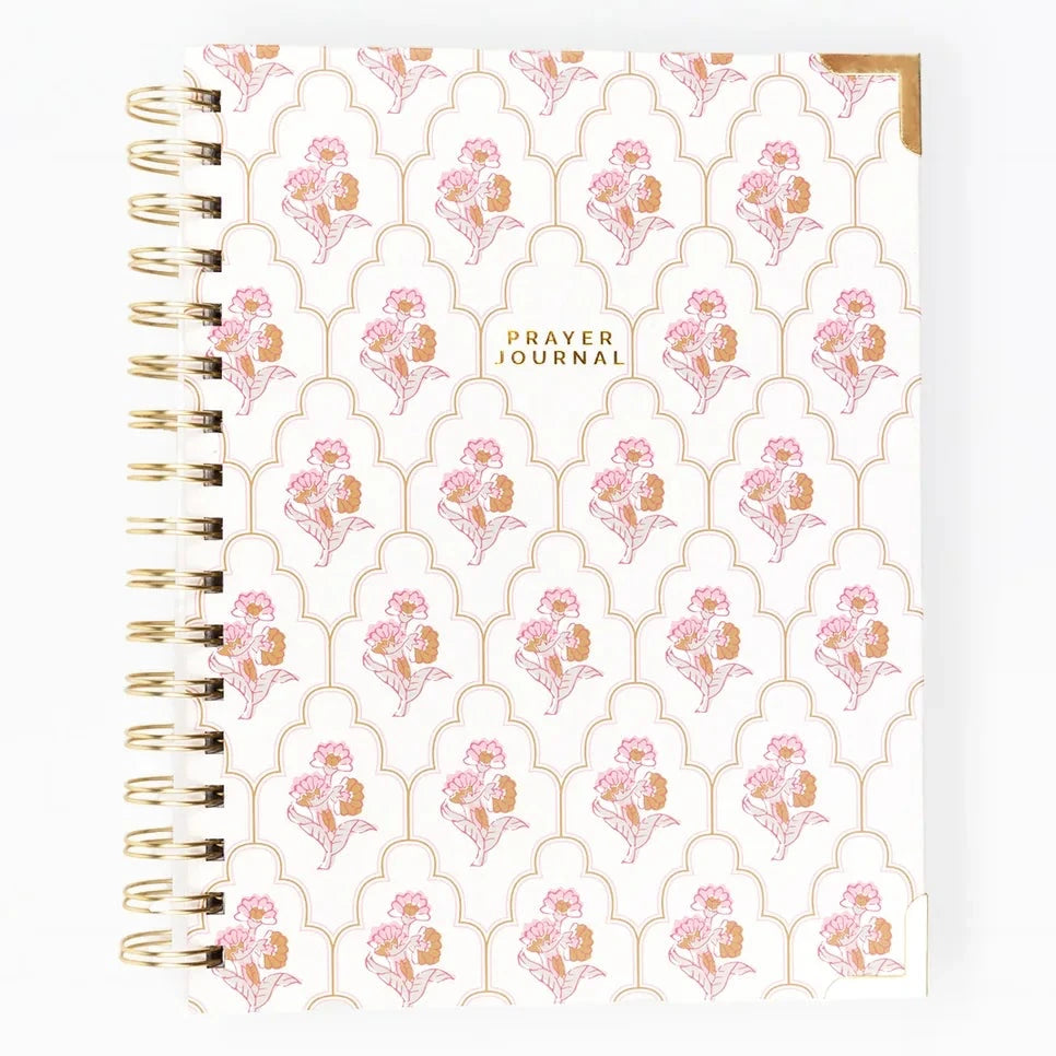 A spiral-bound prayer notebook with a white cover decorated with a pink and gold floral trellis pattern, featuring the text 'PRAYER JOURNAL' in gold near the center.