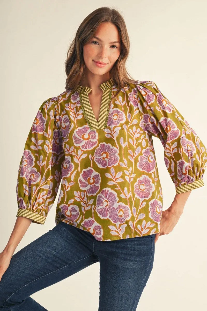 A woman modeling a short-sleeve blouse with a yellow-green base and a purple floral pattern, paired with dark blue jeans, against a plain light beige background.