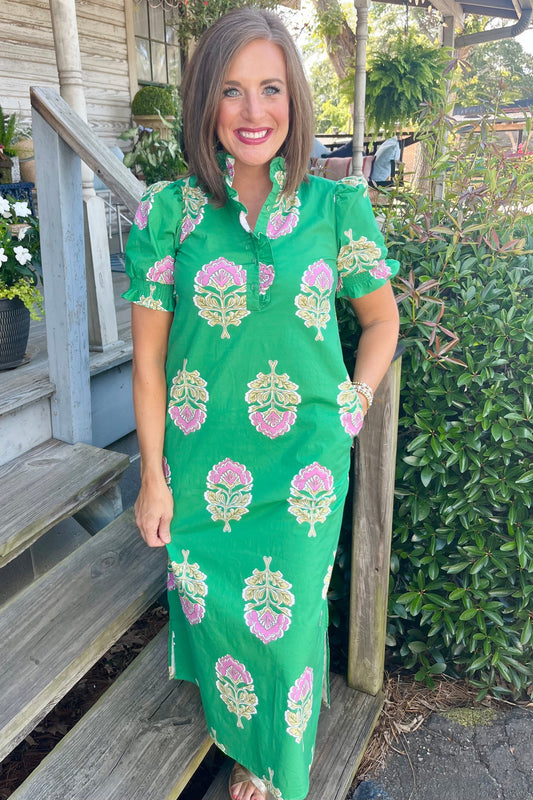 A woman wearing a bright green short-sleeve dress with pink and yellow floral block prints, standing on wooden porch steps outdoors.