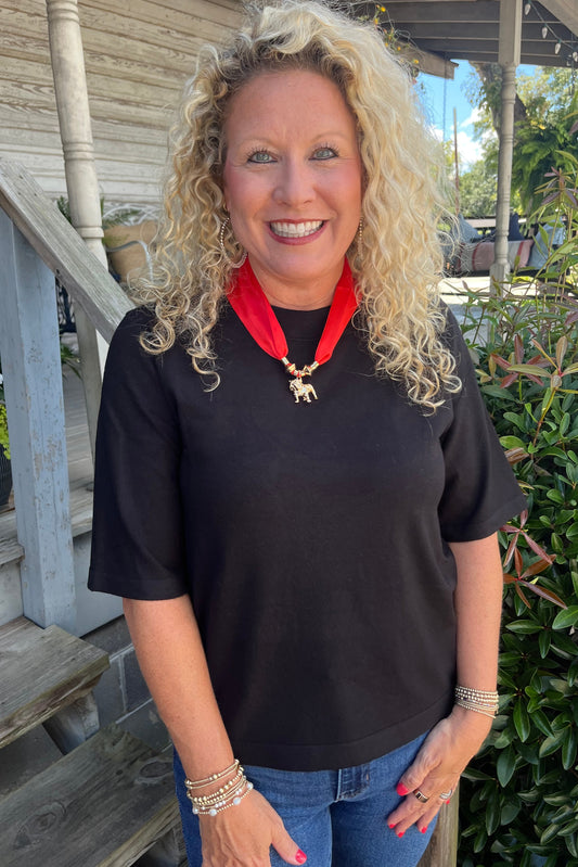 A smiling blonde woman wearing a black short-sleeve t-shirt, blue jeans, and a red neck scarf with a gold elephant pendant, standing on a wooden porch with green plants in the background.