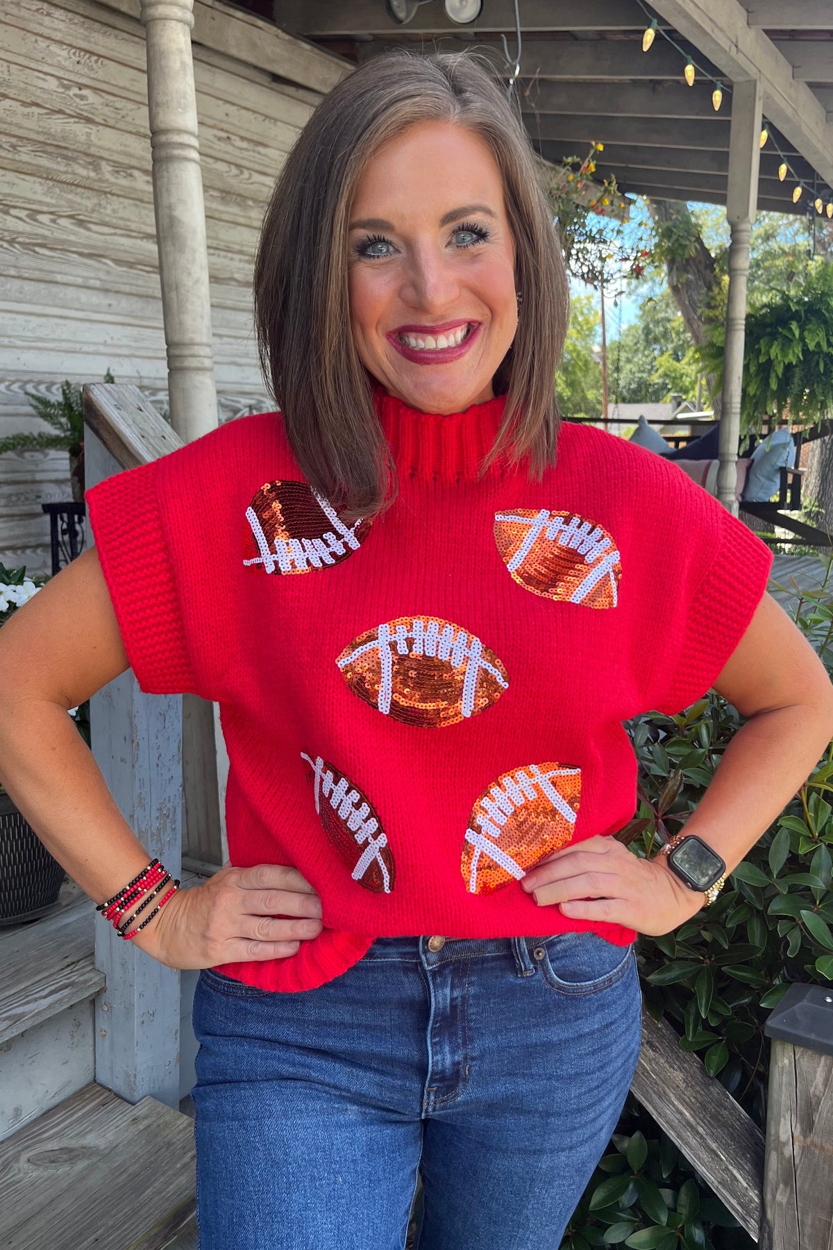 A woman wearing a bright red knit turtleneck sweater with football graphics, paired with blue jeans, standing on a wooden porch.