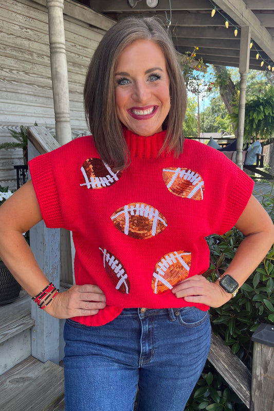 A woman wearing a bright red knit turtleneck sweater with football graphics, paired with blue jeans, standing on a wooden porch.