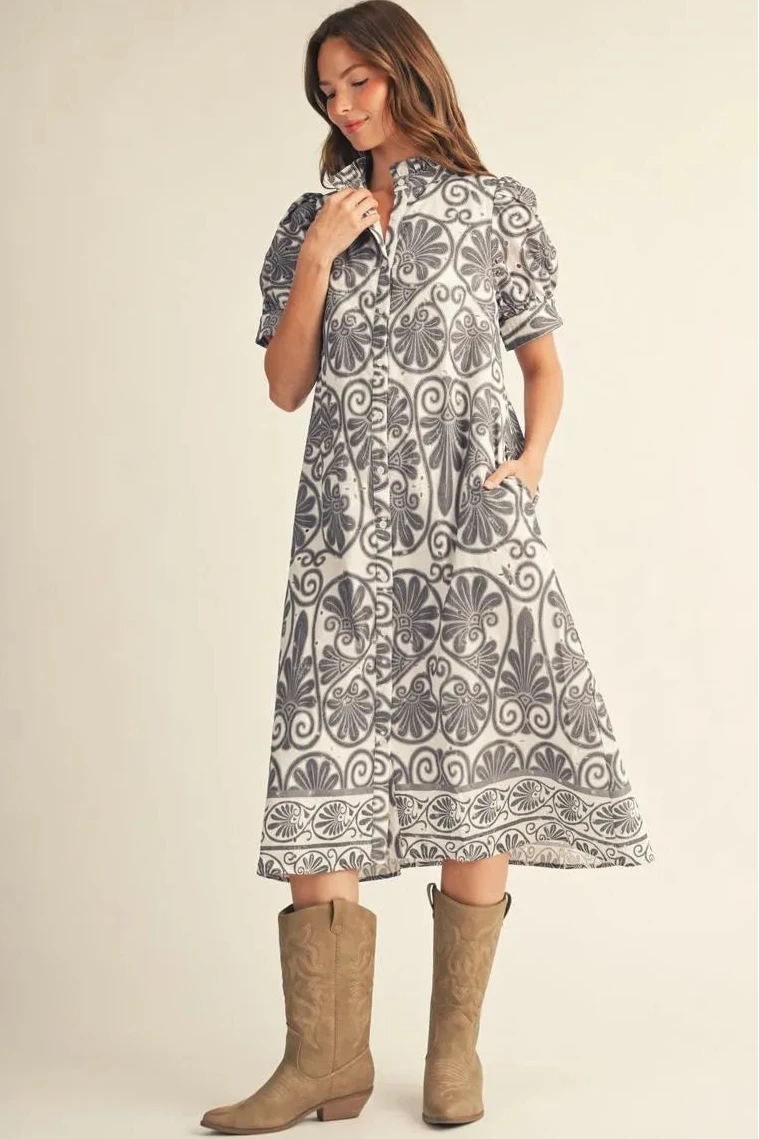 A woman modeling a short-sleeve midi dress with a gray and white floral and scroll pattern, paired with tan cowboy boots, against a solid beige studio background.