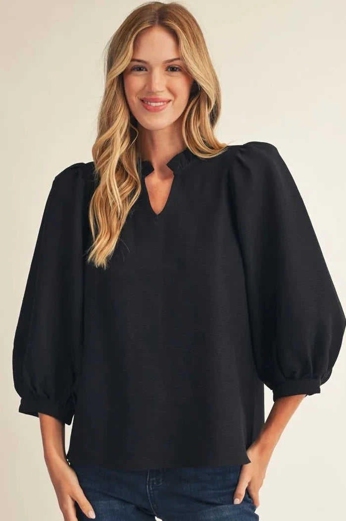 A woman modeling a black long-sleeve blouse with voluminous balloon sleeves and a small keyhole cutout at the neckline, paired with blue jeans against a plain light beige studio background.