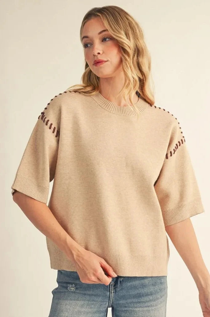 A woman wearing a beige short-sleeve knit sweater with decorative cutout detailing on the shoulders, paired with blue jeans, against a plain light beige background.