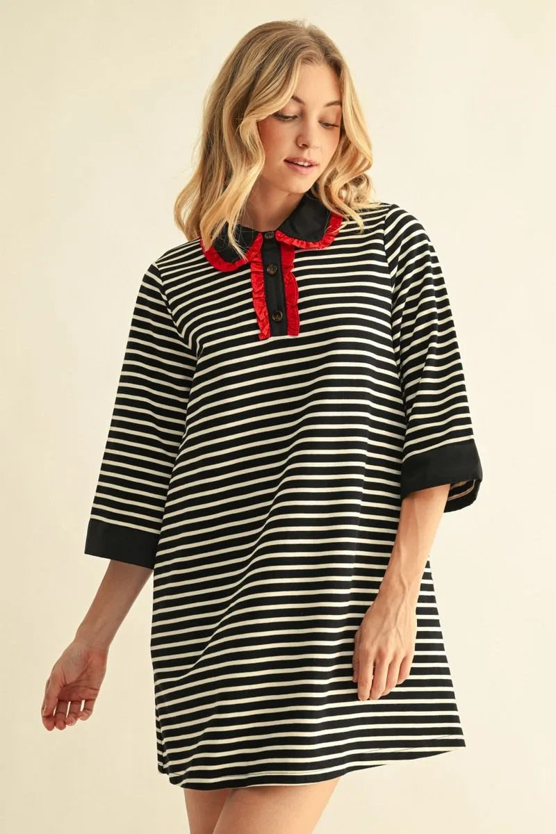 A woman wearing a black and white horizontally striped tunic dress with a red collar and bow detail, photographed against a plain beige studio background.