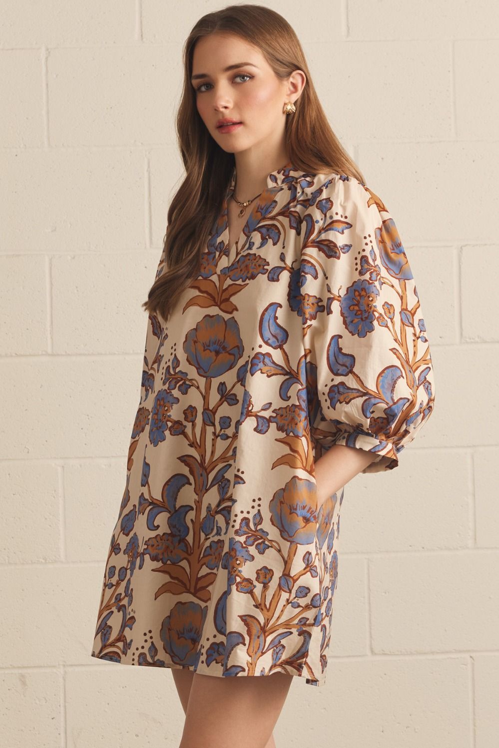 A woman wearing a loose, knee-length dress with a beige base and a blue and brown floral print, standing against a white painted brick wall.