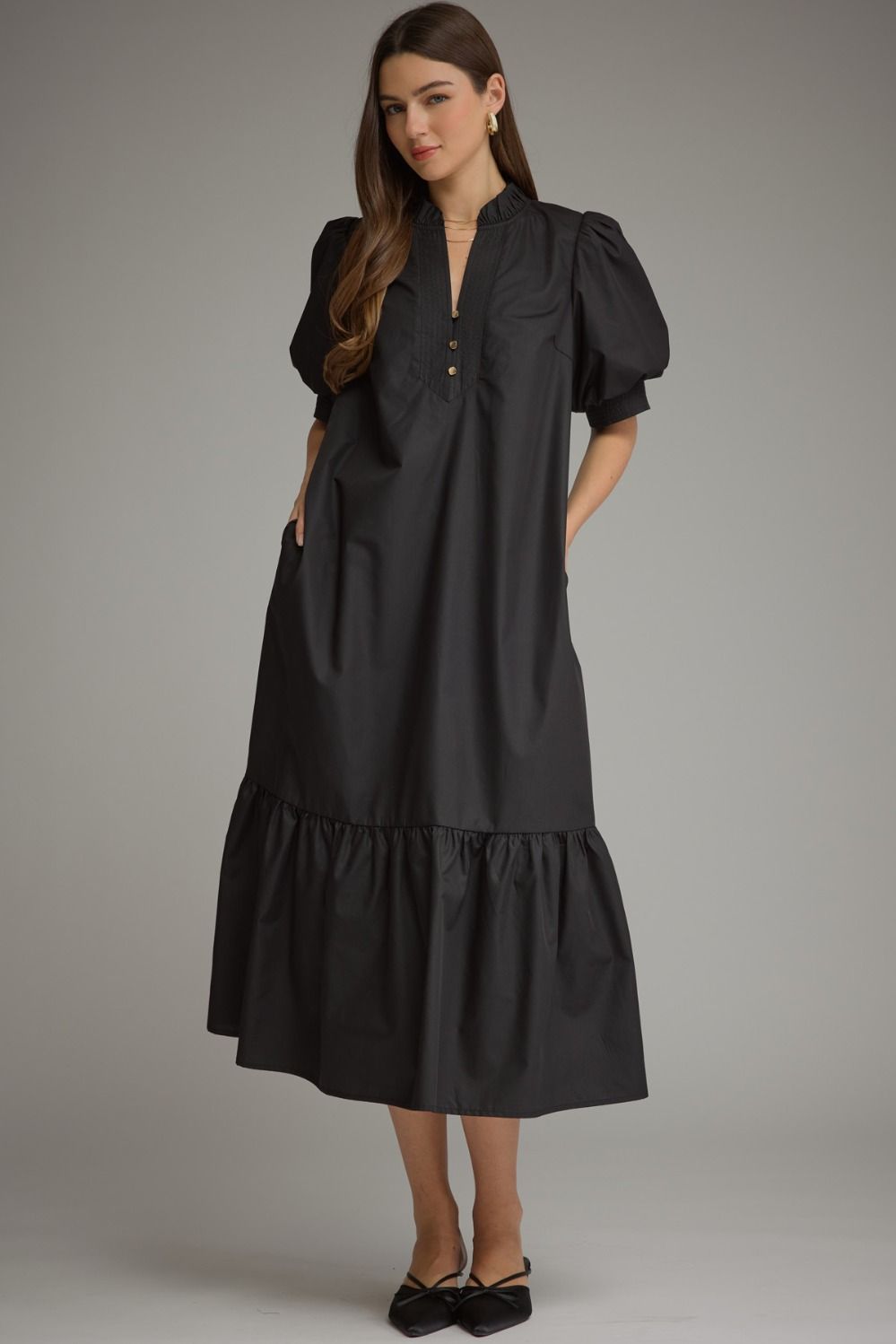 A woman modeling a black midi dress with puff sleeves, a split neckline with decorative gold buttons, and a tiered hem, photographed against a neutral gray studio background.
