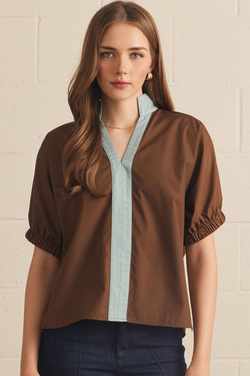 A woman modeling a brown short-sleeve blouse with a light blue chambray V-neck placket and gathered cuffs, paired with dark blue jeans, standing against a light beige brick wall.