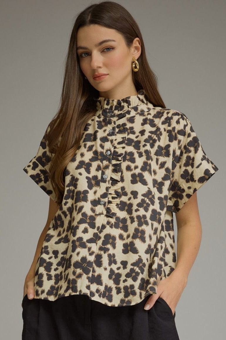 A woman modeling a short-sleeve high-neck blouse with a beige and black leopard print, paired with black shorts, against a solid gray studio background.